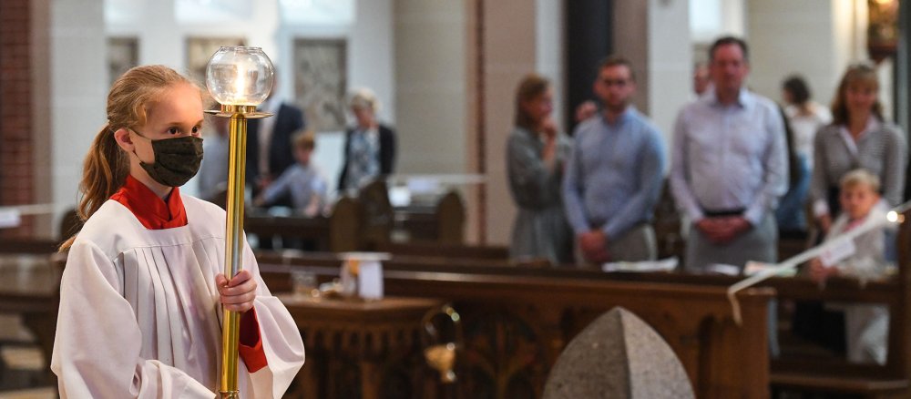 Eine Ministrantin mit Mundschutz hält den Leuchter bei der Messe zur Feier der Erstkommunion am 21. Juni 2020 in einer Kirche in Bonn.