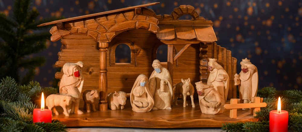 Eine Krippe aus Holz. Alle Krippenfiguren tragen Mundschutz. Neben der Krippe brennen zwei Kerzen.