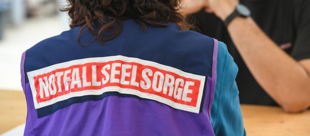 Eine Frau trägt eine Weste mit der Aufschrift "Notfallseelsorge" und spricht mit einem Mann