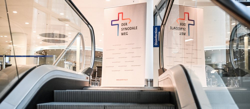 Blick eine Rolltreppe hinauf auf einen Aufsteller mit dem Logo des Synodalen Weges