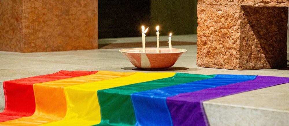 Vier brennende Kerzen stehen in einer Schale neben einer Regenbogenfahne auf den Altarstufen