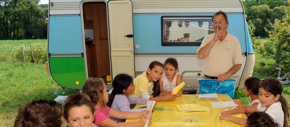 Vor einem Wohnwagen steht ein Pfarrer, er singt mit einigen Kindern.