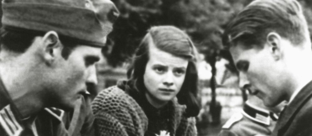 Hans Scholl in Uniform am linken Bildrand, Christoph Probst in zivil am rechten Bildrand. Dazwischen Sophie Scholl mit strengem Seitenscheitel, Strickjacke und ernstem Blick.