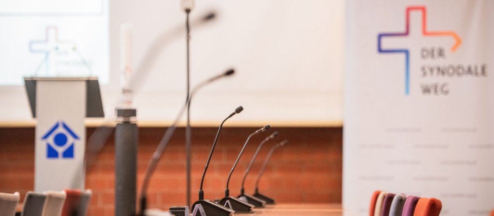 Mikrofone auf Tischen, im Hintergrund das Synodalkreuz und das Logo des Synodalen Weges