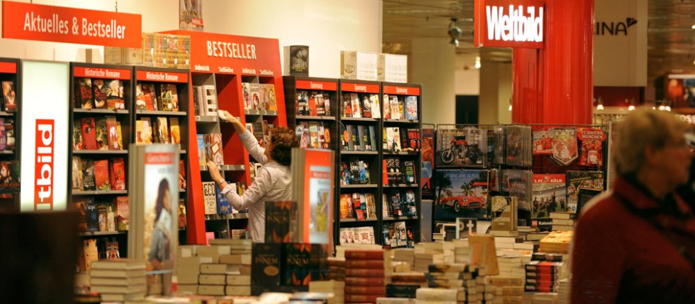 Das Innere einer Buchhandlung: Regale voller Bücher stehen an der Wand, andere Bücher stapeln sich in der Mitte des Raumes, mittendrin stöbern Kunden.