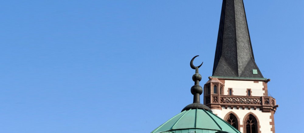 Das Dach der Yavuz Sultan Selim Moschee in Mannheim. Die Moschee liegt in unmittelbarer Nachbarschaft zur katholische Liebfrauenkirche. Minarett und Kirchturm ragen in den blauen Himmel.