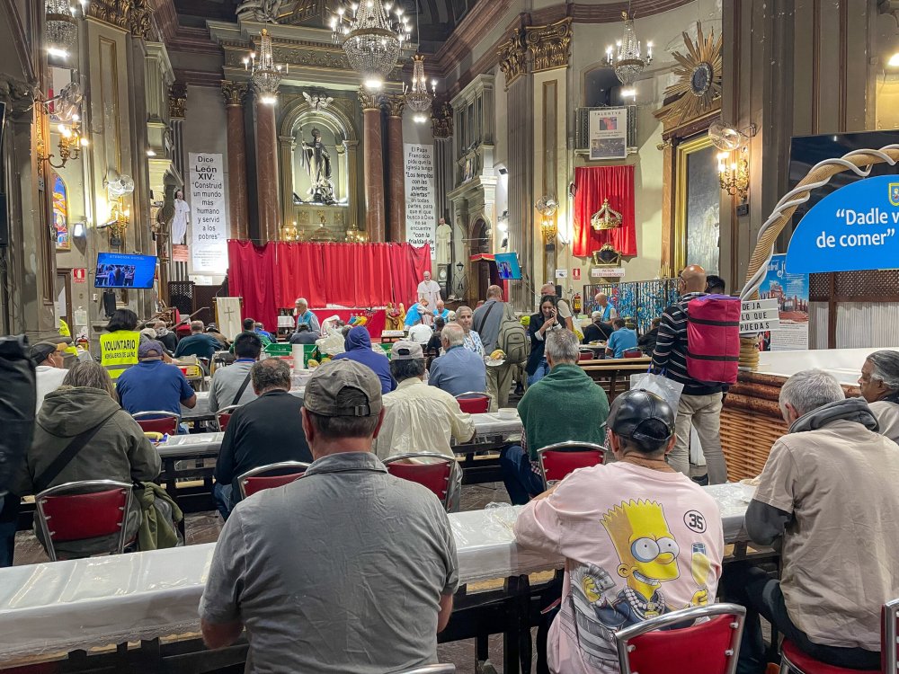 Viele bedürftige Menschen sitzen auf roten Stühlen an Tischreihen in einer Kirche und essen, den Blick Richtung Altar.
