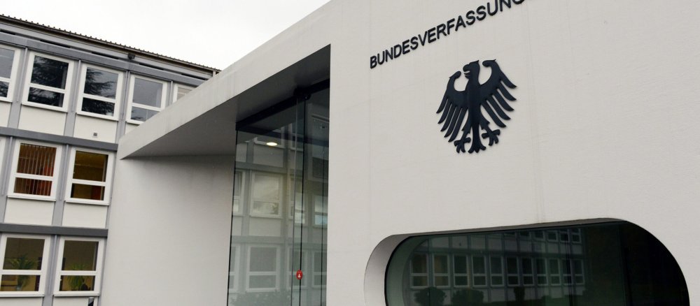 Der Tür eines weißens viereckigen Hauses. Daneben ein großes ovales Fenster über dem der Bundesadler und die Aufschrift "Bundesverfassungsgericht" angebracht ist.