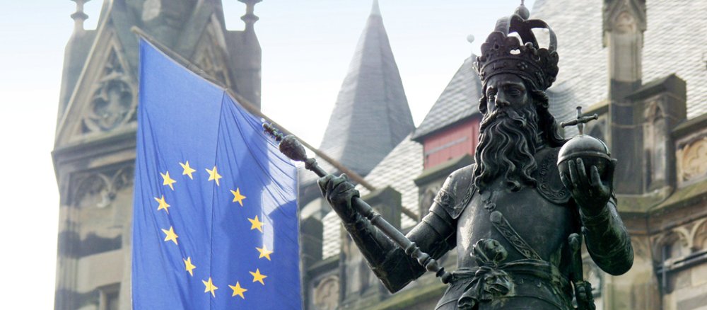 Die Bronzestatue zeigt einen Mann mit Vollbart, die Kaiserkrone auf dem Kopf, Zepter in der rechten, den Reichsapfel in der linken Hand. Im Hintergrund weht die EU-Fahne.