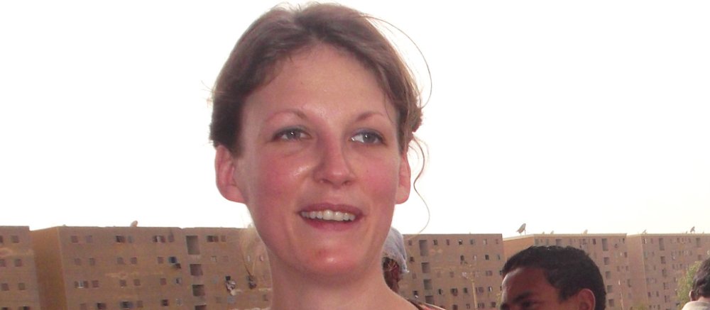 Ein brünette Frau, bekleidet mit einer weißen Bluse und einer roten Weste, steht vor einer Häuserfront.