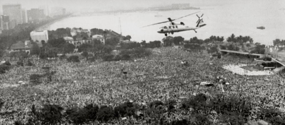 Auf dem Foto sieht man eine große Menschenmasse von mehreren Millionen Menschen auf engem Raum. Über ihnen schwebt ein Hubschrauber mit Papst Johannes Paul II. an Bord. Im Hintergrund sind Häuser von Manila und die Küste zu sehen.