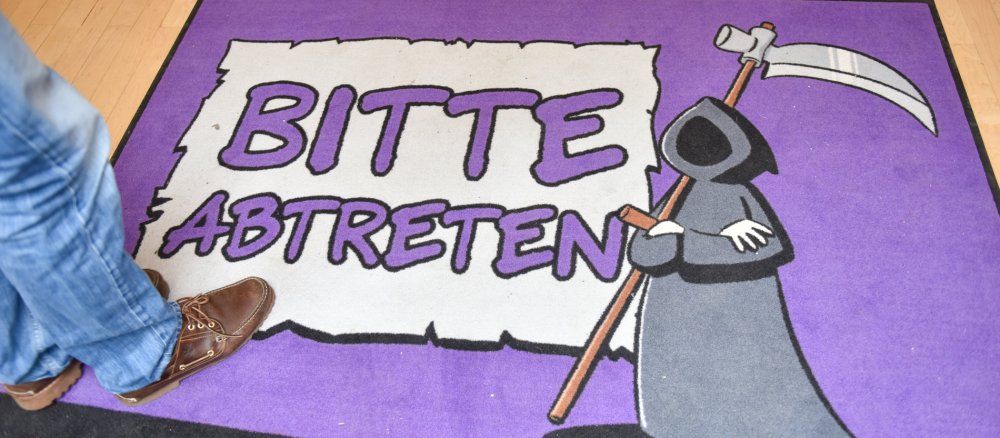 Eine Fußmatte, auf der der Sensenmann abgebildet ist. Daneben steht: "Bitte abtreten."