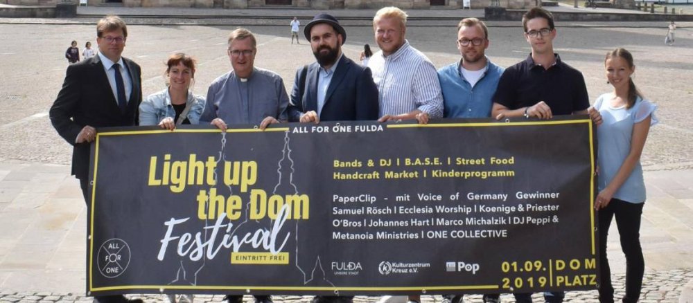 Acht Personen, die Veranstalter des Light up the Dom Festivals in Fulda, halten ein großes Werbeplakat des Festival in die Höhe.
