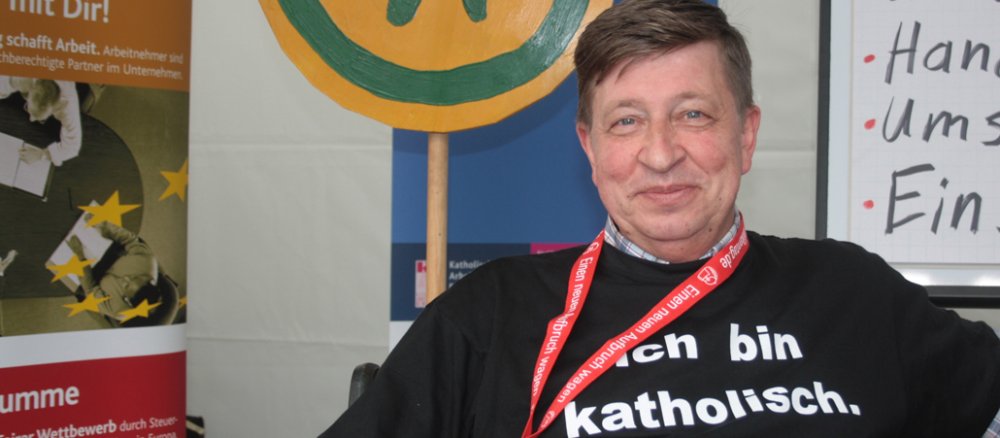 Norbert Faltin sitzt im schwarzen T-Shirt vor einigen Schildern. Auf seinem T-Shirt steht "Ich bin Katholike" geschrieben.