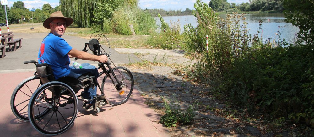 Ein Rollstuhlfahrer steht mit seinem Handbike an einem See