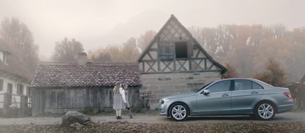 Vor einem Bauernhof stehen zwei Mädchen beim Spielen, neben ihnen steht ein Mercedes