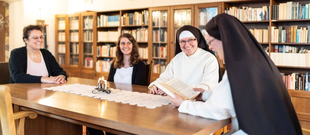 Zwei Frauen und zwei Ordensfrauen sitzen an einem Tisch, eine der Ordensfrauen liest etwas vor.