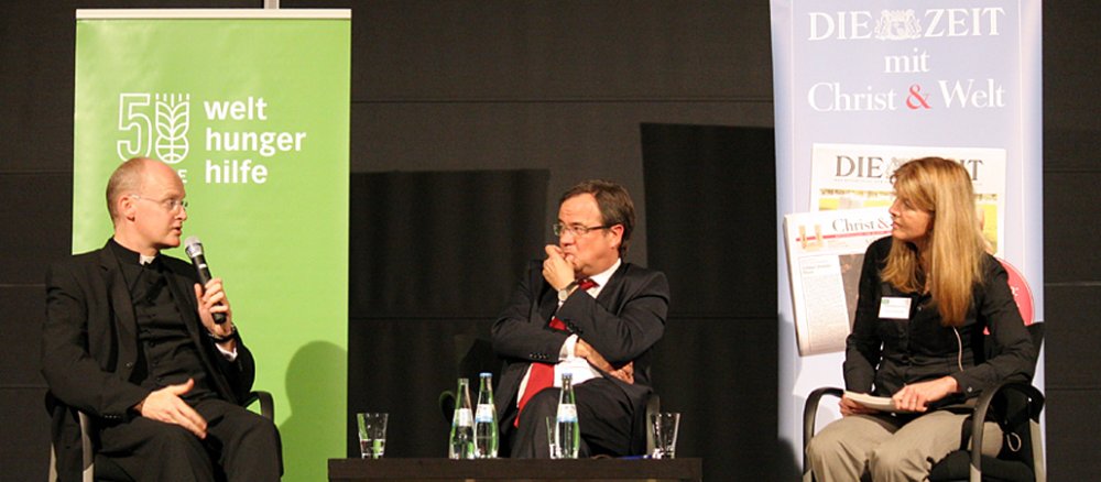 Bischof Franz-Josef Overbeck (links) zusammen mit Armin Laschet (mitte) und Moderatorin Astrid Prange de Oliveira auf der Podiumsdiskussion "Wem gehört die Welt?".
